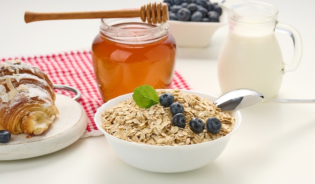 Desayuno matutino, copos de avena cruda en un plato de cerámica, leche en un decantador, arándanos y miel en un tarro sobre una mesa blanca