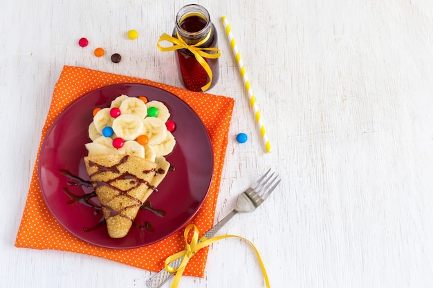 Desayuno o postre para niños: tortitas con plátano, cobertura de chocolate y caramelos de colores. Elaboración de alimentos dulces en forma de helado.