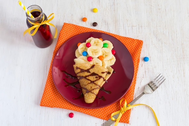 Desayuno o postre para niños: tortitas con plátano, cobertura de chocolate y caramelos de colores. Elaboración de alimentos dulces en forma de helado.