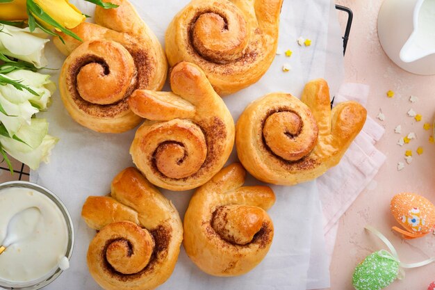 Desayuno de Pascua Concepto de vacaciones Rollos de bollos de conejito de Pascua con canela hechos de masa de levadura con glaseado de naranja decoraciones de Pascua huevos de colores sobre fondo rosa de primavera Tarjeta de vacaciones de Pascua