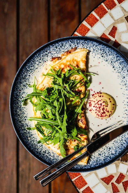 Un desayuno que consiste en una tortilla con rúcula o ensalada de rúcula con salsa en una mesa de piedra de mosaico de un café en un día soleado, vista superior