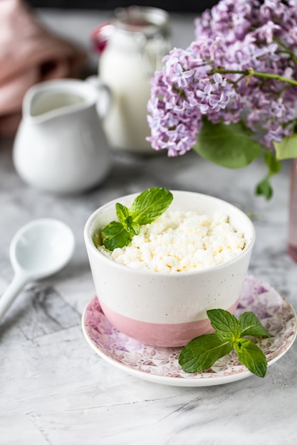 Desayuno requesón con arándanos, crema, leche sobre una mesa blanca y una rama de flores.