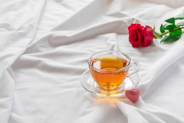 Desayuno romántico en la cama con una taza de té, dulces de color rosa en forma de corazón y rosa sobre sábanas blancas.