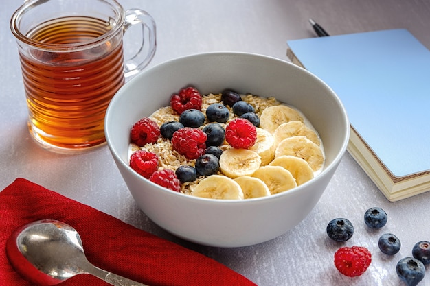 Desayuno saludable con avena en un bol, rodajas de plátano, frambuesa, arándano, una taza de té y un cuaderno con un bolígrafo sobre la mesa gris claro