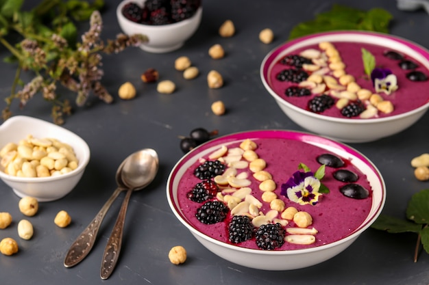 Desayuno saludable batido tazón con moras, grosellas, maní, avellanas, decorado con flores sobre un fondo oscuro