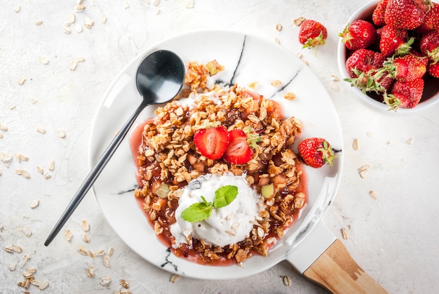 Desayuno saludable. Crumble de avena y granola con ruibarbo, fresas frescas y moras, semillas y helado en placa de mármol blanco, con menta, en mesa de hormigón de piedra blanca, vista superior de copyspace