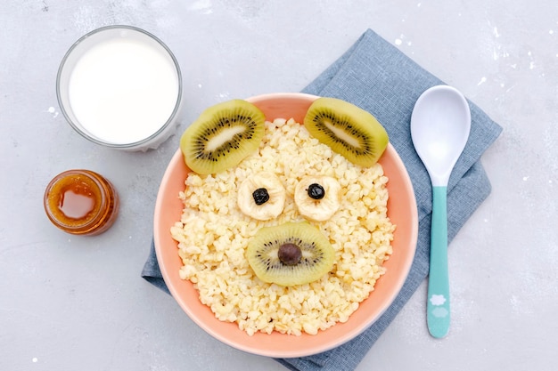 El desayuno saludable para el desayuno del bebé de los niños lindos y divertidos para niños en un tazón parece una cara de ratón de oso decorada con kiwi, plátano, bayas secas, frutas, leche, postre, arte de comida en una mesa gris