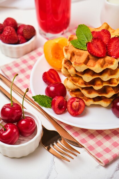 Desayuno saludable con gofres belgas con fresas, albaricoques, cerezas, jugo y una taza de café negro y chocolate amargo sobre fondo de mesa de piedra blanca