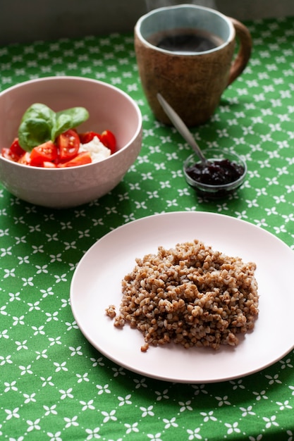 desayuno saludable y nutritivo: un rico plato de trigo sarraceno, una ensalada de requesón y tomate con albahaca, un tazón pequeño de mermelada de ciruela pasa, una taza de café caliente sobre un mantel verde