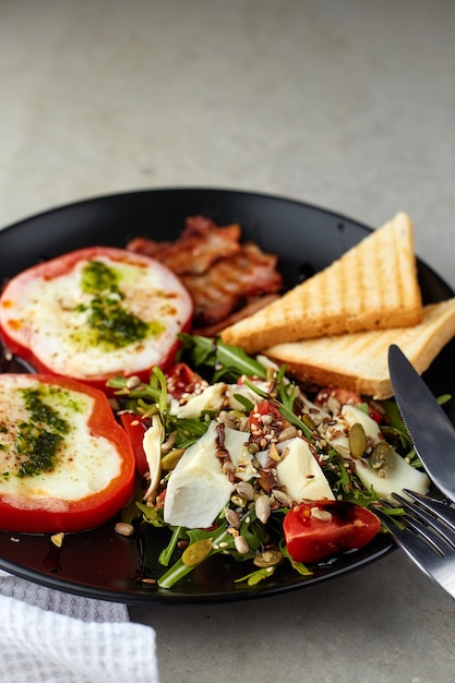 Desayuno saludable y sabroso. Huevos fritos en aros de pimiento rojo, tocino y ensalada con rúcula, tomates cherry, semillas y queso duro en placa negra