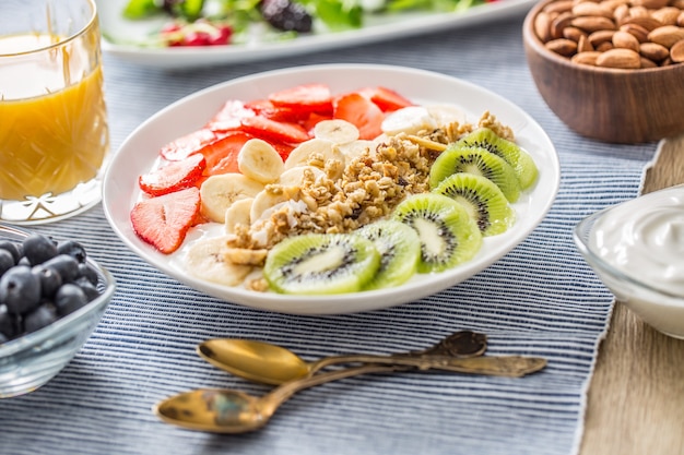 Desayuno saludable servido con plato de yogur, muesli, kiwi, fresas y plátano. Mesa de la mañana granola, almendras, jugo de bayas y hierbas verdes.