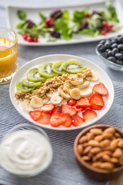 Desayuno saludable servido con plato de yogur, muesli, kiwi, fresas y plátano. Mesa de la mañana granola, almendras, jugo de bayas y hierbas verdes.