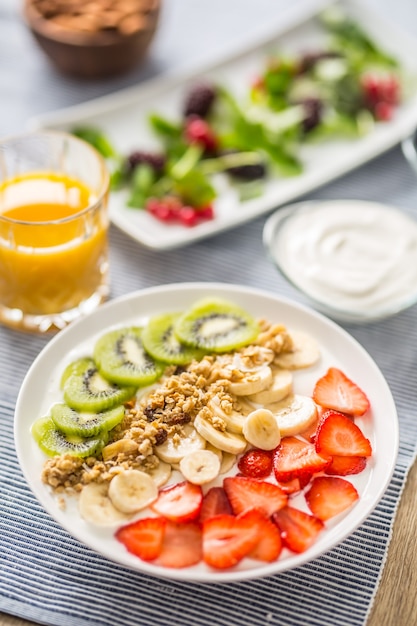 Desayuno saludable servido con plato de yogur, muesli, kiwi, fresas y plátano. Mesa de la mañana granola, almendras, jugo de bayas y hierbas verdes.