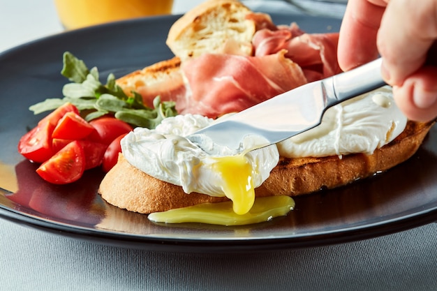 Desayuno saludable con tostadas de pan integral y huevo escalfado. La mano del hombre con un cuchillo cortando un huevo