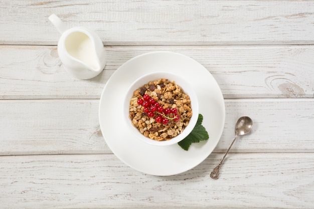 Desayuno saludable de verano de granola, muesli con jarra de leche con decoración de grosella roja en tablero de madera. Vista superior.