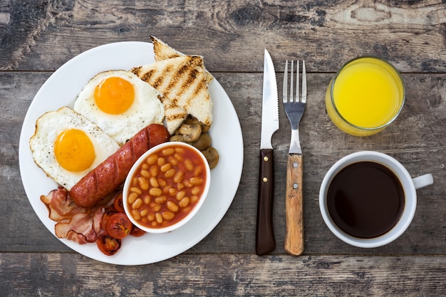 Desayuno tradicional inglés completo con huevos fritos, salchichas, frijoles, champiñones, tomates asados y tocino sobre superficie de madera