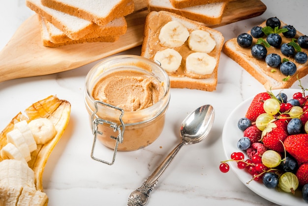 Desayuno tradicional de verano americano y europeo: sándwiches de pan tostado con mantequilla de maní, bayas, frutas manzana, durazno, arándano, arándano, fresa, plátano. Mesa de mármol blanco. copyspace