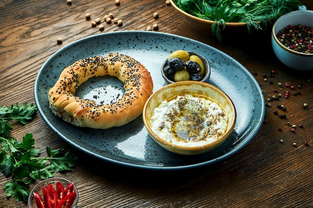 Desayuno turco clásico: simit con mousse de queso feta con aceite de oliva y especias, servido en un plato azul con aceitunas. Mesa de madera. Comida de restaurante. Simit - bagel con semillas de amapola o semillas de sésamo