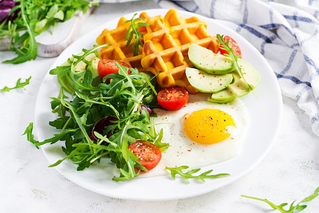 Desayuno con waffles de calabaza, huevo frito, tomate, aguacate y rúcula sobre superficie blanca