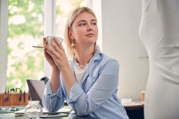 Descansada mujer caucásica mirando a otro lado y inclinando los codos sobre la mesa mientras toma una taza de café