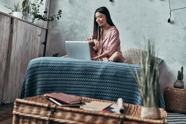 Descansando en casa. Mujer joven atractiva que usa la computadora y sonriendo mientras está sentado en la cama en casa