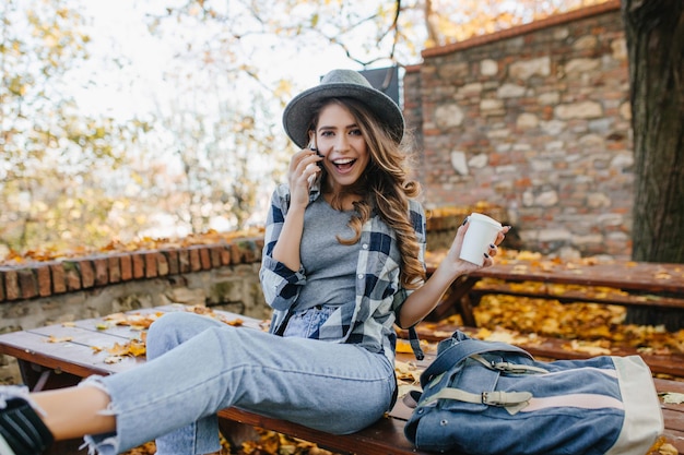 Descansando dama blanca en jeans casuales cortos llamando a alguien sobre fondo de otoño. Retrato al aire libre de la chica europea de moda de buen humor disfrutando de un café en el día de noviembre.