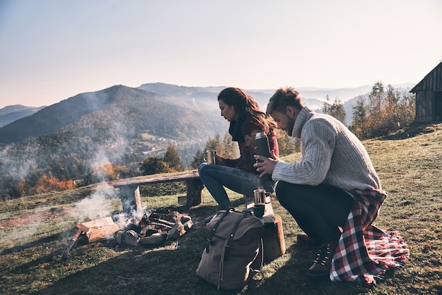 Descansando juntos. Lindo casal jovem tomando café da manhã enquanto está sentado perto da fogueira nas montanhas