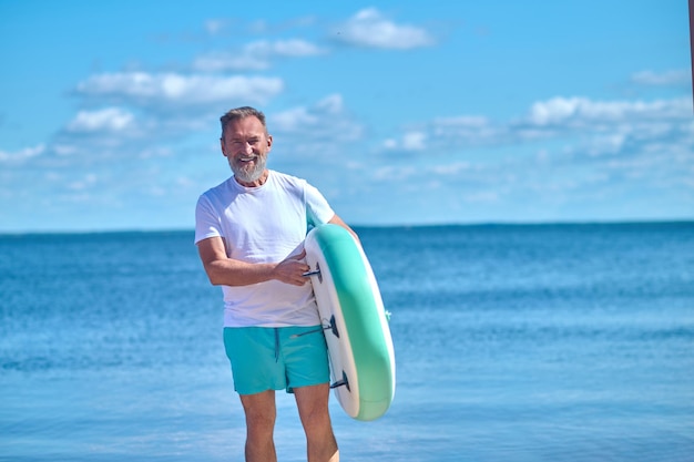 Descanse, mar. Homem barbudo grisalho em pé com paddleboard inflável sorrindo para a câmera na beira-mar em dia ensolarado