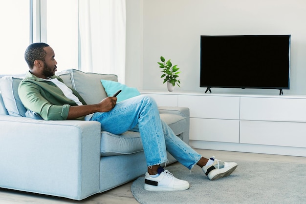 Descanse y relájese Hombre negro sentado en el sofá en la sala de estar viendo un programa de televisión mirando televisión de plasma en blanco