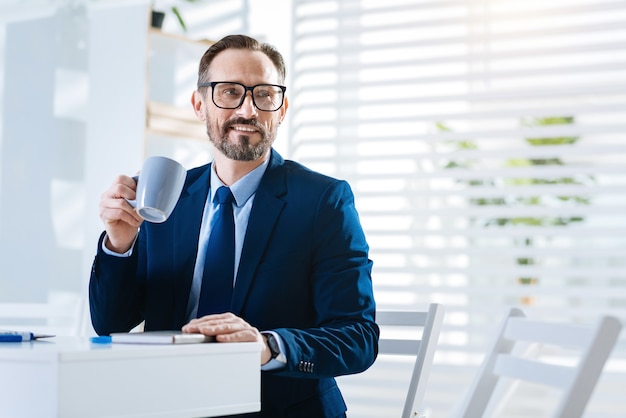 Descanso. Feliz atractivo hombre vigoroso cuidando café mientras está sentado en la mesa y sonriendo