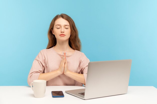 Descanso en el trabajo, práctica de yoga. Una empleada tranquila sentada en el lugar de trabajo con una laptop y levantando las manos en un gesto de namaste, meditando en la oficina de casa. Foto de estudio interior aislado sobre fondo azul.