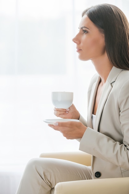 Descanso. Vista lateral de la pensativa joven empresaria en traje tomando café y mirando a otro lado mientras está sentado en la silla