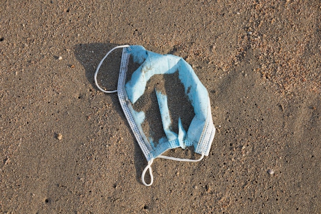 Descartado na praia do mar máscara facial azul pálida de uso único Poluição ambiental e costeira Máscara cirúrgica usada jogada na praia ameaçando a saúde do oceano Resíduos durante o COVID19