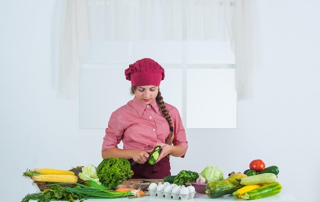 Descasque uma menina adolescente de pepino preparando refeição vegetais crus frescos hora de comer criança feliz cozinhando na cozinha alimentos saudáveis e orgânicos apenas cheios de vitaminas criança usar uniforme de chef e chapéu