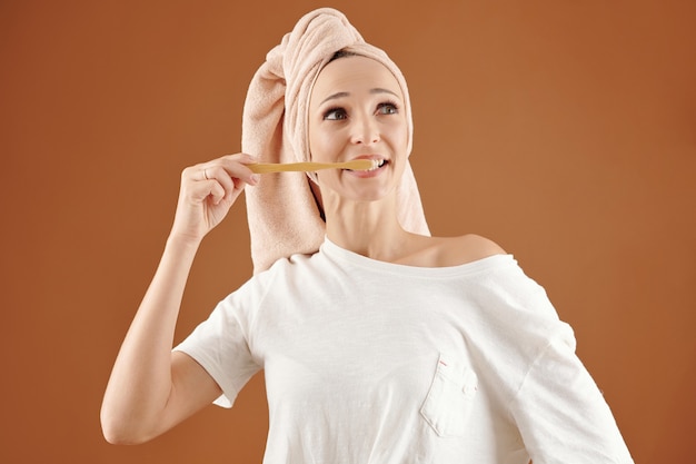 Desconcertado joven mujer caucásica con el pelo envuelto en una toalla de baño cepillarse los dientes contra el fondo marrón