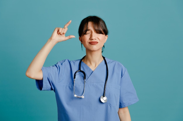 Descontente mostrando tamanho jovem médica vestindo estetoscópio uniforme de quinto isolado em fundo azul