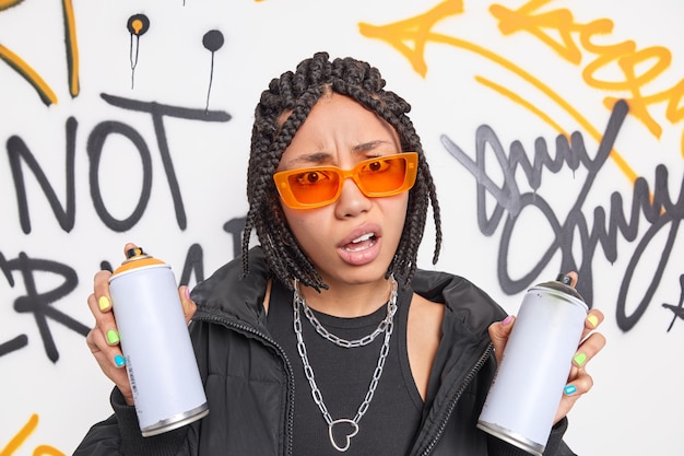 Descontento adolescente indignado con rastas sonríe cara sostiene aerosol no le gusta algo dibuja graffiti en lugar urbano usa gafas de sol naranjas de moda