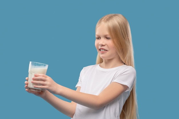 Descontento. Chica de escuela primaria rubia disgustada en camiseta blanca empujando vaso de leche