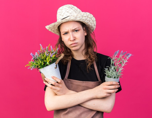 Descontento joven jardinero vistiendo sombrero de jardinería sosteniendo y cruzando flores en macetas