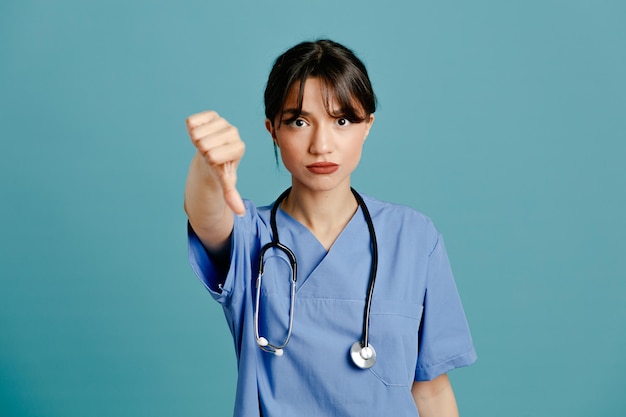 Descontento mostrando los pulgares hacia abajo joven doctora vistiendo uniforme fith estetoscopio aislado sobre fondo azul.