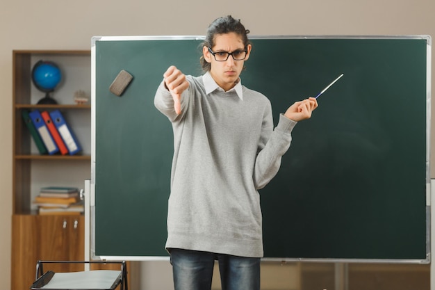 descontento mostrando los pulgares hacia abajo joven maestro parado frente a la pizarra y apuntando a la pizarra con el puntero en el aula