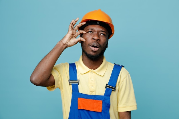 Descontento poniendo la mano en la cara joven constructor afroamericano en uniforme aislado sobre fondo azul.