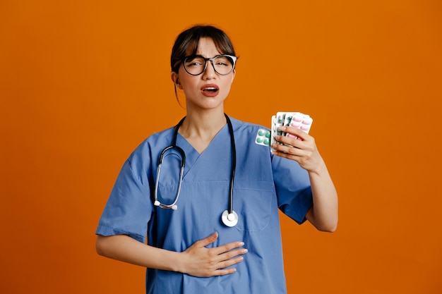 Descontento poniendo la mano en el estómago sosteniendo píldoras joven doctora vistiendo uniforme fith estetoscopio aislado sobre fondo naranja