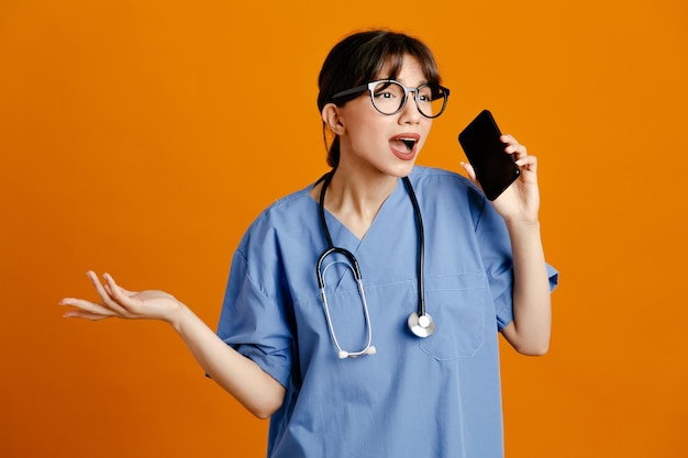 Descontento sosteniendo teléfono joven doctora vistiendo uniforme fith estetoscopio aislado sobre fondo naranja