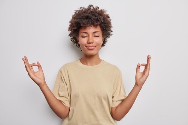 Descontraído e tranquilo jovem de cabelos cacheados faz gesto zen mantém hads em mudra fecha os olhos pratica ioga usa camiseta casual isolada sobre fundo branco se acalma durante dia estressante