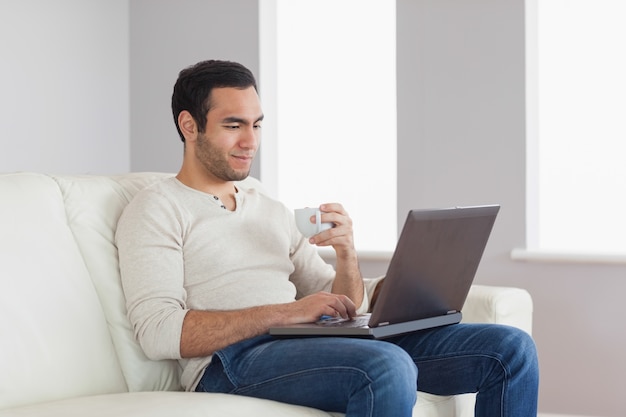 Descontraído homem atraente segurando café enquanto trabalha em seu laptop