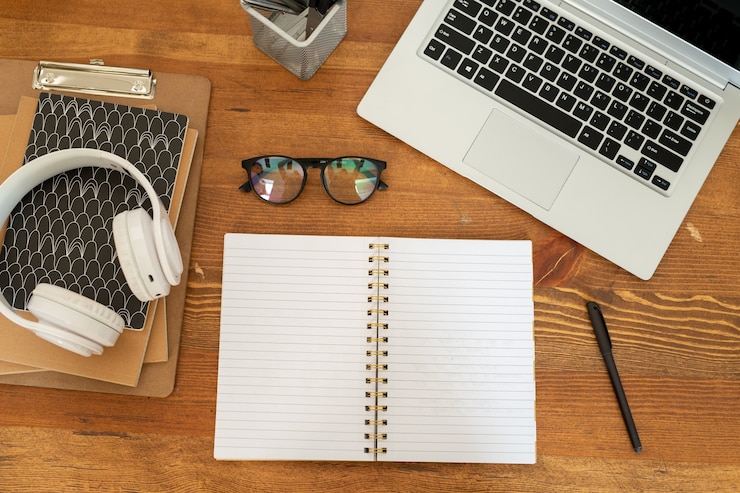 Foto descripción general de cuaderno abierto con páginas en blanco rodeadas de portátil, anteojos, auriculares, blocs de notas y bolígrafo