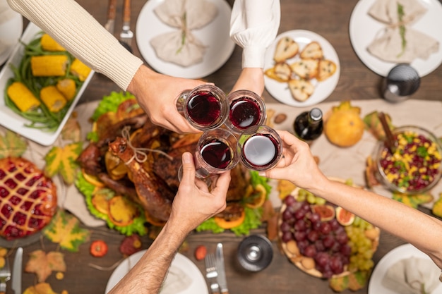 Descripción general de cuatro copas de vino tinto sostenidas por miembros de la familia tintineando sobre una mesa festiva servida con pavo casero y otros alimentos