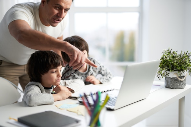 Descubra la mejor clase. Padre cariñoso que ayuda a sus hijos durante la lección en línea para niños. Niños latinos mirando la pantalla del portátil, sentados juntos en la mesa en casa