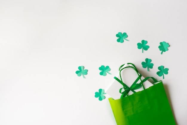 Descuentos para el día de San Patricio. Bolsa de papel verde y hojas de trébol de papel, caja de regalo con un lazo verde sobre un fondo blanco.